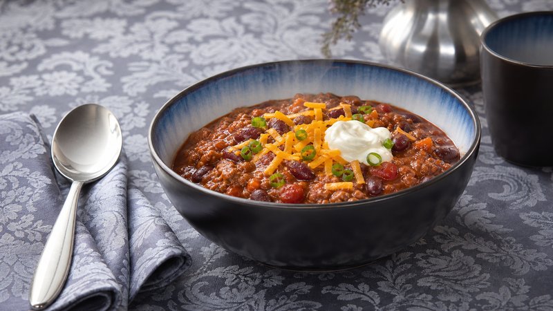 A steaming bowl of classic beef chili topped with shredded cheddar cheese and sour cream, with tortilla chips on the side