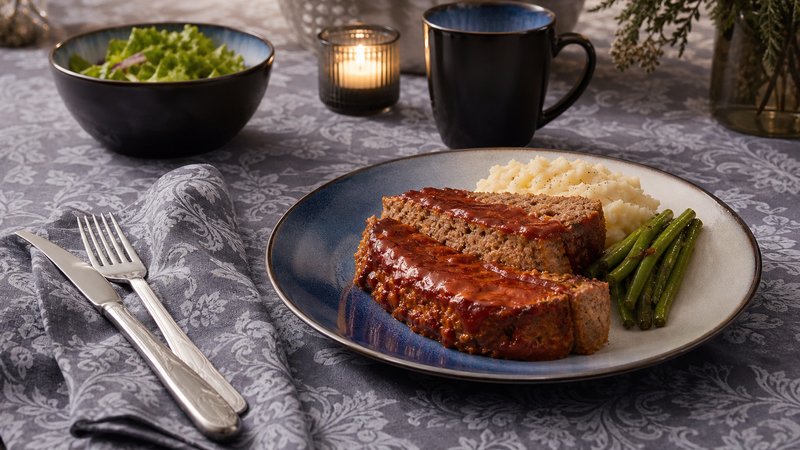 A thick slice of classic meatloaf with glazed ketchup-brown sugar topping, served on a plate with creamy mashed potatoes and green beans