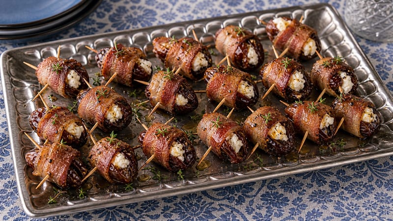 Bacon-wrapped dates stuffed with goat cheese arranged on a white serving platter, golden brown and glistening, garnished with fresh thyme