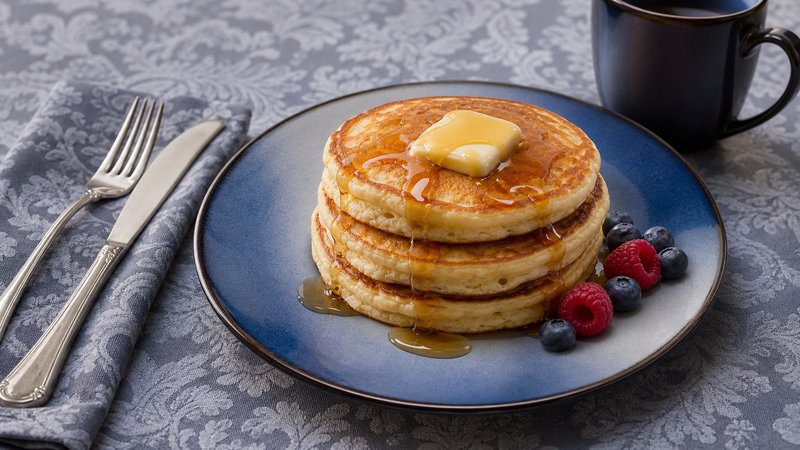 A stack of golden-brown fluffy pancakes topped with fresh strawberries, whipped cream, and a drizzle of maple syrup