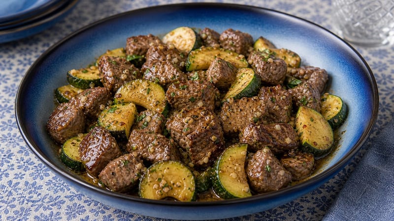 Cast iron skillet of golden-seared garlic butter steak bites plated alongside caramelized zucchini, garnished with fresh parsley on a dark wood surface
