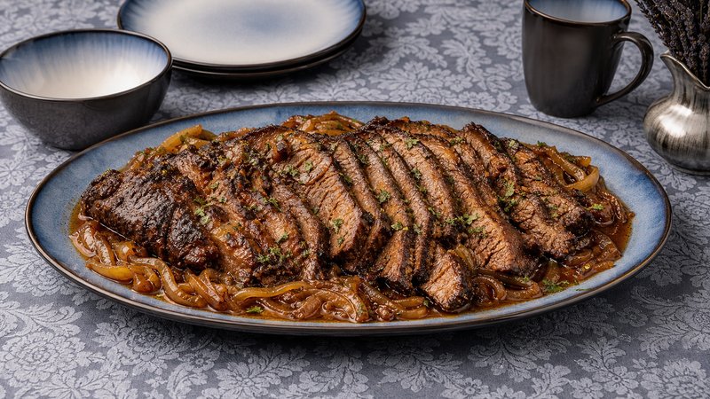 Sliced oven-baked beef brisket with onions and pan sauce, served on a plate with roasted vegetables
