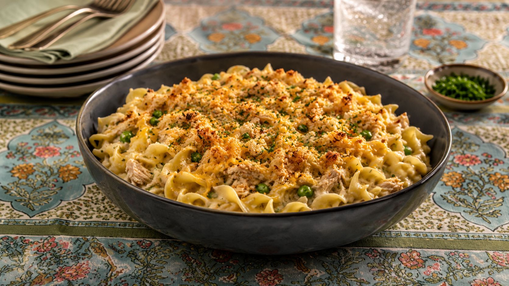 A golden tuna noodle casserole in a white baking dish, topped with crispy buttered breadcrumbs and garnished with fresh parsley, with a serving spoon lifted to show the creamy interior